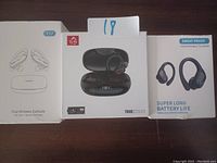 Three boxes of wireless sports earbuds lined up on a dark surface. Box 1 shows True Wireless Earbuds with simple line drawing and charging case. Box 2 is open showing black earbuds inside charging case with a digital display on the case. Box 3 shows an image of black earbuds with ear hooks and claims of 'Sweat Proof' and 'Super Long Battery Life'.