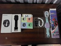 Photo of five fitness trackers and smart watches arranged on a wooden surface including boxes, charging cable, and plastic packaging.