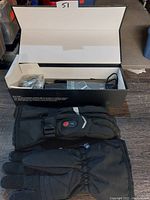 Box open showing black Savior heated gloves with control button on the back of glove, power adapter and user guide inside box, and partially visible exterior storage bag.