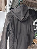 Back view of black heated jacket with hood hanging on rack, showing material and design.