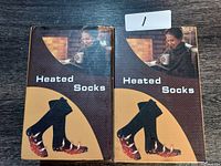 Photo of two boxes of heated socks on a dark wood surface, one box marked with a label '1'.