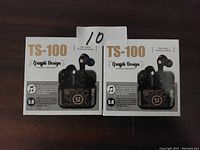 Two boxed TS100 wireless stereo earbuds with graphite design shown front facing on a dark surface, labeled with number 10.