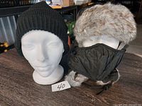 Photo showing a front view of both hats on white mannequin heads, the black knit beanie and the faux fur trapper hat with ear flaps and attached black mask.