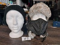 Two winter hats displayed on mannequin heads: a black knit hat and a black quilted trapper hat with faux fur.