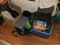 Wide angle showing various bags in a blue plastic basket and surrounding area including grey and black backpacks and a colorful lunchbox