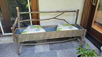 Front view of rustic wooden bench with natural tree branch frame, showing galvanized planter area and two removable cushioned seats with green clover and blue floral pattern.