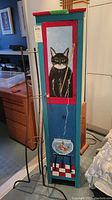 Front view of tall wooden cabinet painted teal with red border and black and white cat artwork holding a fishing pole with fishbowl painted on door.