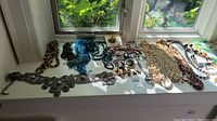 Overview of multiple necklaces, pendants, and metal belt arranged on a white surface near a window. Shows diversity of materials including beads, shells, stones, and metal.