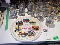 Photo showing eight tall glasses with black and gold Montreal street scenes and white round Expo 67 souvenir tray.