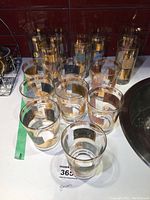 Full set of 15 drinking glasses arranged on a surface near a sink, showing eight shorter glasses in front and seven taller glasses behind, all having gold rim and stamp designs on glass surface.