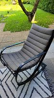 Side view of black metal framed outdoor rocker chair with gray padded cushion seating on patterned outdoor rug and stone patio.
