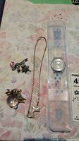 Photo showing Swatch watch in clear plastic case alongside rose brooch, bird pendant necklace, and bunny brooch on floral surface