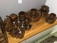 Overall view of all pieces on wooden shelf: lidded pots, pitchers, stacks of plates, cups and mugs.