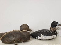 Two wooden duck decoys, one brown and one black and white, resting side by side with visible strings tied around their necks.