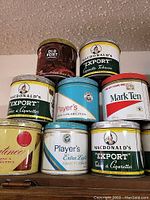 Photo showing eight different vintage cigarette and tobacco tins stacked in two rows, featuring brands such as MacDonald's Export, Player's, Mark Ten, Old Port, and Valentine.