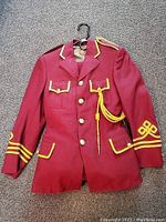 Front view of maroon band uniform jacket with yellow trim, gold buttons, multiple pockets and shoulder cord hanger on black hanger.