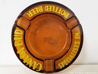 Amber glass ashtray with text 'CANADIAN BUD', 'BOTTLED BEER', and 'SPECIALISTS IN' in yellow around edges. Four cigarette rests spaced evenly.