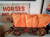 Photo showing the covered wagon light with an orange fabric cover and wooden wheels against a backdrop including a vintage style wooden sign that reads 'HORSES NOT ALLOWED ON'.