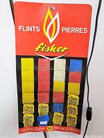 Metal store display sign holding multiple colored packs of Fisher flints, with red top section bearing Fisher logo and flame design.
