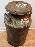 Front view of rusty metal milk can with lid and handle, showing worn and faded paint markings.