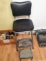 Front view of retro stool chair showing black cushioned seat and backrest, metal legs, and two metal footrests.