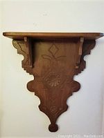 Frontal view of the small wooden wall shelf showing carved front panel and support brackets beneath the shelf.