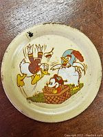 Photo showing the round metal tip tray with vintage Donald Duck and hen illustration on a cream background, with visible age wear and some rust spots.