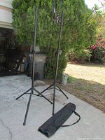 Pair of black adjustable PA speaker/lighting stands with tripod bases set up outdoors on concrete surface next to greenery. A black zippered carrying bag lies flat on the ground in front of the stands.