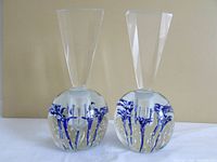 Pair of clear round art glass perfume bottles with blue flower motif inside glass. Tall faceted clear glass stoppers and bubble inclusions in the glass. Bottles sit side by side on a flat surface.