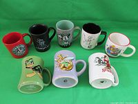 Full view of all eight mugs arranged on green background showing various colors and character designs from Disney, DC Comics, and Warner Bros.