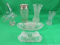 Photo showing six cut glass items arranged side by side on a green background. Items include three vases, two trays and one basket with handle. The glass is clear with intricate cut designs.