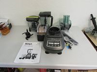 Full view of Ninja blender and food processor components arranged on table including motor base, blender pitcher, food processor bowl, discs, blades, power cord, and manual.