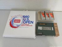 Photo showing three sleeves of orange Wilson ProStaff golf balls, a box of 10 Spalding Professional II golf balls, and a 1980 US Open Tennis Championships seat cushion with sponsor logo.