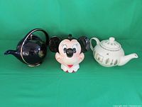 Three teapots side by side on green background: dark Pfaltzgraff Naturewood teapot, Minnie Mouse shaped teapot missing bow, cream colored Hall Pottery teapot with botanical decals.