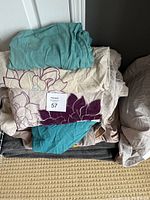 Stack of assorted bedding including floral duvet cover with purple and cream colors, teal-green sheet, and folded bedding items below