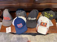 Six baseball caps arranged on a bench showing front logos including Under Armour, Fox, Spider, Easton, Canada Archery, and one cap with yellow text.