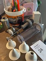 Photo of various cake icing nozzles, clear press-style icing dispenser, metal cone tip, and plastic kitchen bucket with utensils inside on table surface