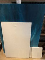 Large painted canvas (36x48) with blue and teal abstract swirling pattern visible, medium white blank canvas (24x36), and one small white melamine board in front showing marks and dents.
