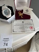 White Casio Baby-G wristwatch in original tin, Camrose & Kross hula girl brooch in burgundy box, certificate of authenticity laid out on white surface