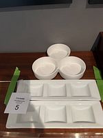 Photo showing three white round ceramic bowls and two white rectangular divided serving trays on a wooden surface.