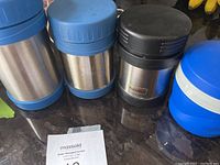 Four thermoses shown side by side on a dark countertop: two with blue lids and bases, one with black lid and Thermos brand, and one blue plastic with embossed logo.