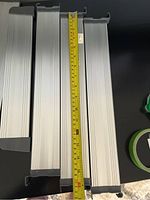 Seven silver aluminum drawer dividers with black plastic ends arranged on black background with measuring tape next to some pieces showing lengths about 20-22 inches.