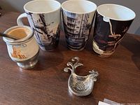 Three tall mugs with cityscape designs, Argentinian tea steeper with metal lid and straw, and metal chicken-shaped tea bag holder arranged on table.