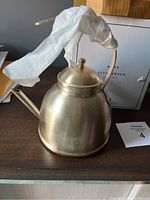 Full view of the tea kettle showing its brushed gold finish and shape with the original box in the background.