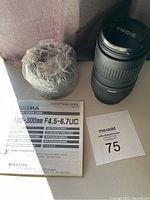 Photo showing Sigma 100-300mm zoom lens, user manual, lens hood wrapped in plastic and auction tag
