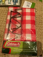Photo showing Coleman picnic tablecloth in red and white checkered design with metal clamps inside packaging.