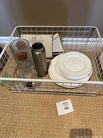 Photo showing wire metal basket containing white plastic plates, clear glassware, stainless steel water bottle without lid, and Pampered Chef jar opener.
