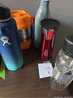 Photo showing all five beverage containers on a table: Hydro Flask, Bubba Keg, stainless steel bottle, Contigo travel mug, and clear plastic bottle.