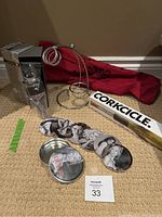 Kuraidori wine decanter aerator in black and grey packaging, boxed and sealed. Corkcicle wine chiller in white and yellow box. Guess coasters set of 6 with black/white female face designs in a round metal Guess branded tin. Metal wine decanter rack with circular holders. Red table runner fabric rolled visible.