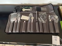Wide-angle photo showing all 11 kitchen utensils neatly arranged in a black rollup organizer, with gray rubber handles. Includes graters, peelers, mashing tools, whisks, and spoons.
